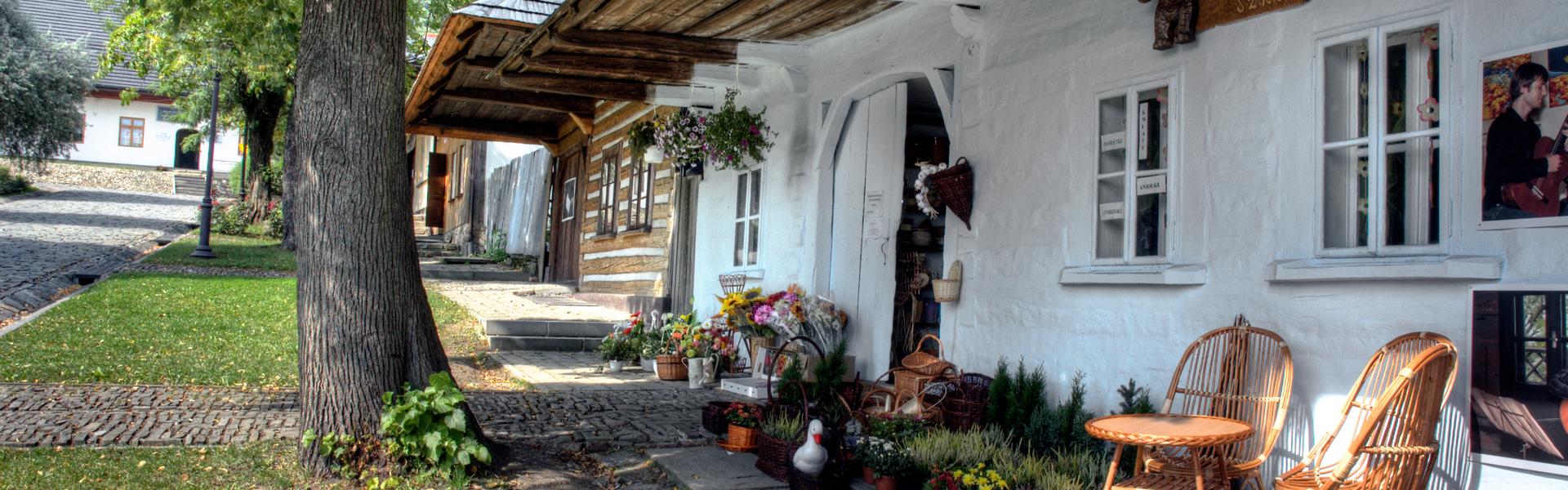 Fragment budynku pomalowanego na biało. Pod ścianą postawione dwa drewniane krzesła. Wokół budynku płot, na którym znajdują się kwiaty w doniczkach.