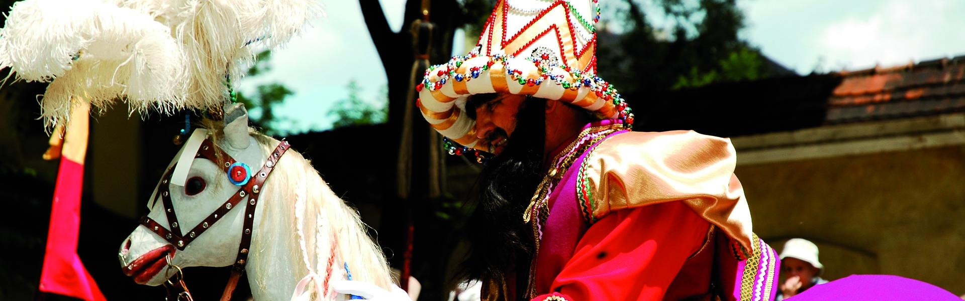 Lajkonik mit dem erhobenen Feldherrnstab in bunter, asiatisch anmutender Kleidung auf dem Krakauer Hauptmarkt. Im Hintergrund Menschen und Türme der gotischen Marienkirche.