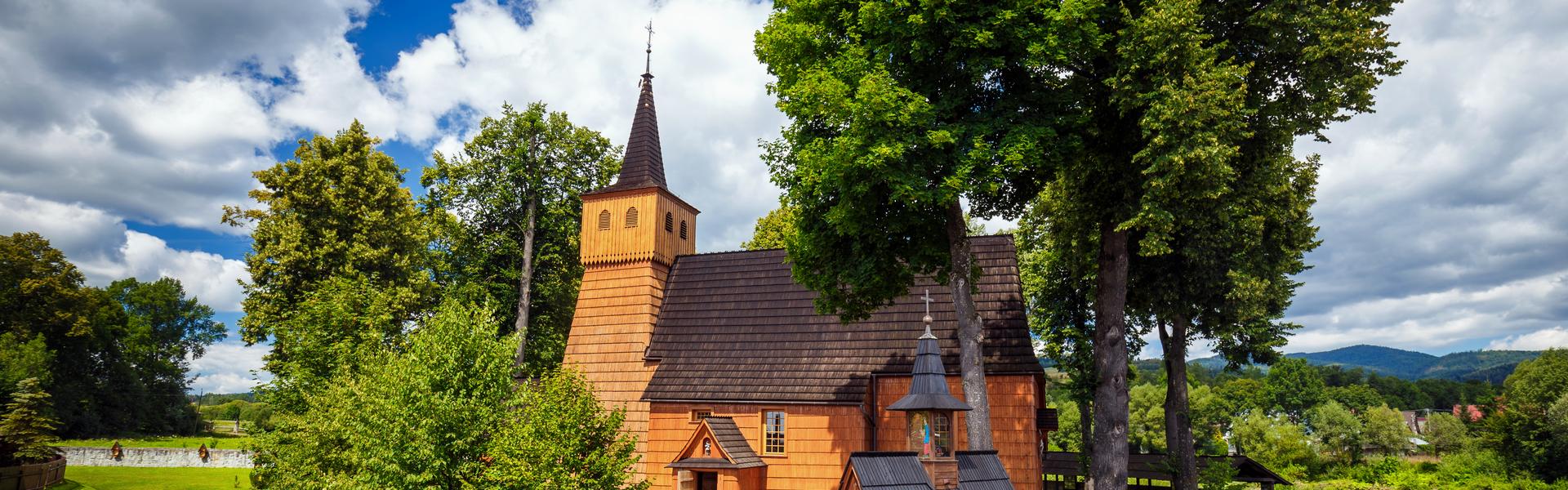 Drewniany kościół o jasnych ścianach i ciemnym dachu, z wieżą. Przy nim kilka drzew oraz drewniane ogrodzenie na kamiennej podmurówce. W tle widać góry.