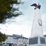 Wysoki kamienny obelisk z orłem na szczycie i krzyżem w wieńcu laurowym. Z tyłu zabudowania.